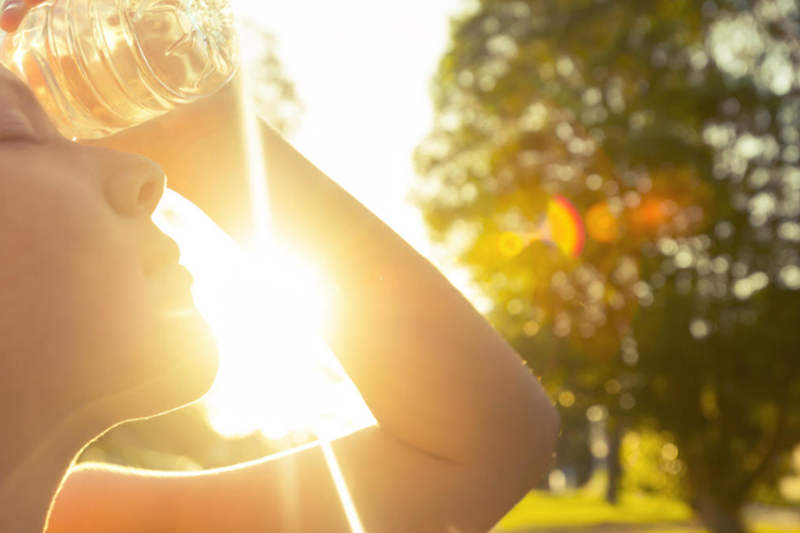 Prepare for Hot Weather With an Air Conditioning Tune-up. Photo of a woman outside using a water bottle to cool off with the sun in the background.